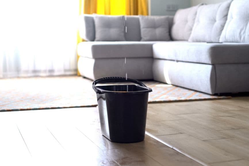 A black bucket catching water dripping from ceiling near a gray sofa and yellow curtains