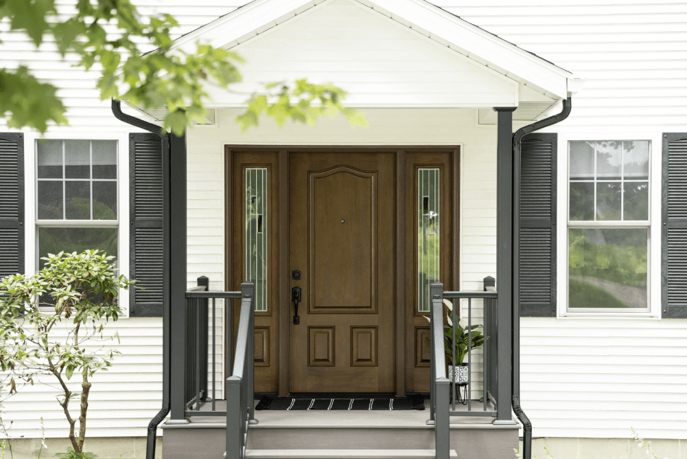 Front view of a white house with a wooden double door, black shutters, and small porch with plants and railing