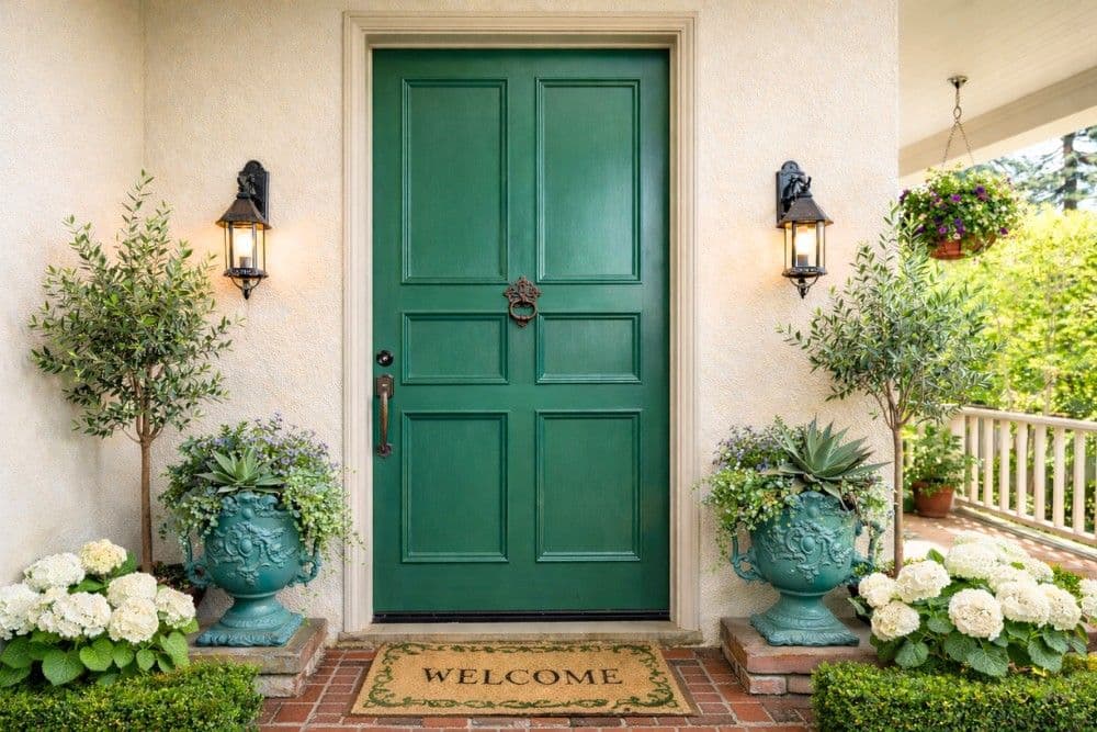 Green six-panel front door with lanterns, urns, and welcome mat