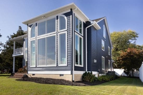 Modern blue house with large energy-efficient windows, green lawn, and trees under a clear sky